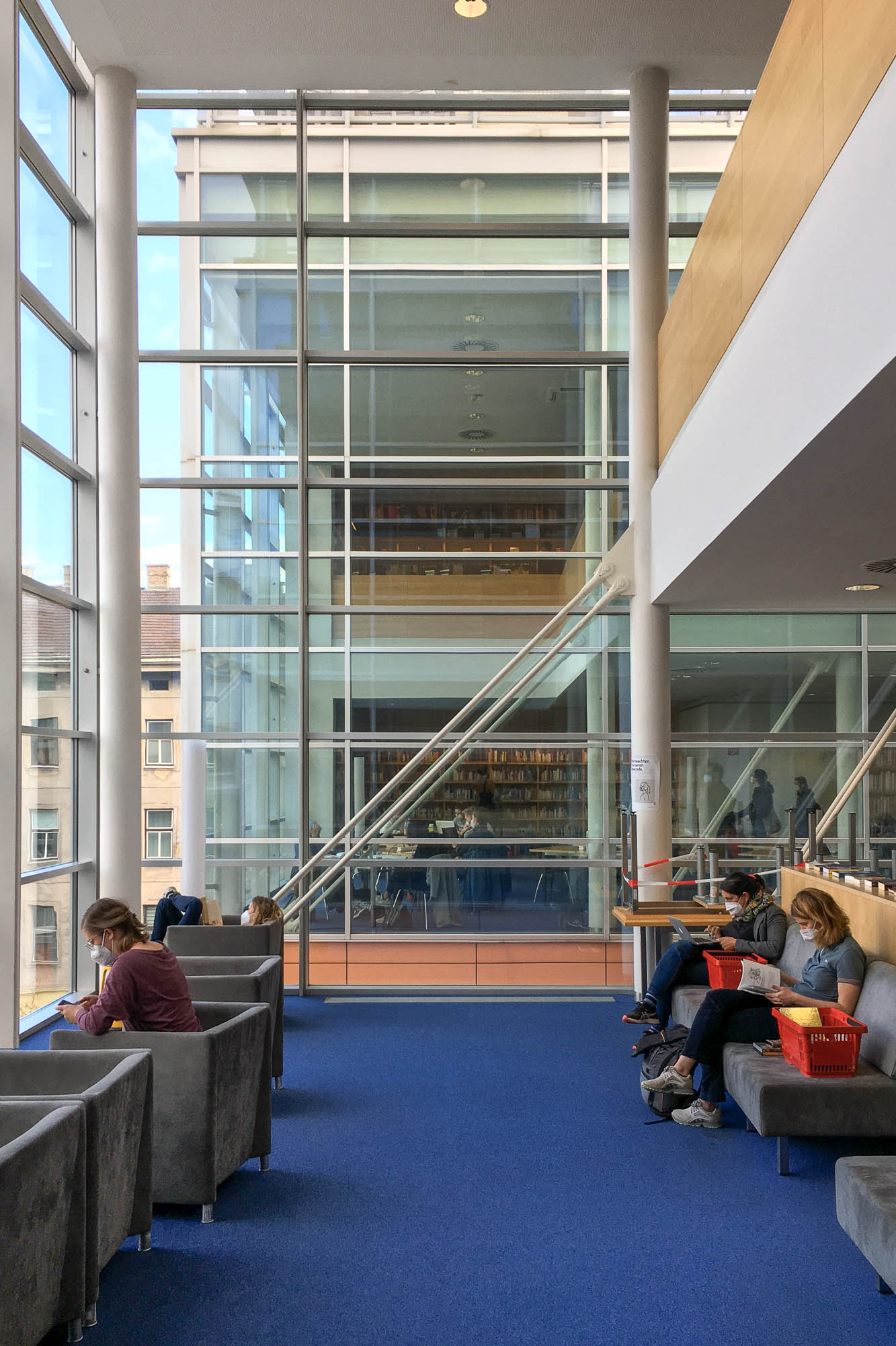 Interior view of a contemporary library. It's a double-height glass-enclosed room with blue carpeting and patrons wearing face masks sitting on couches, reading. Another similar room can be seen across a light well in front of the camera.