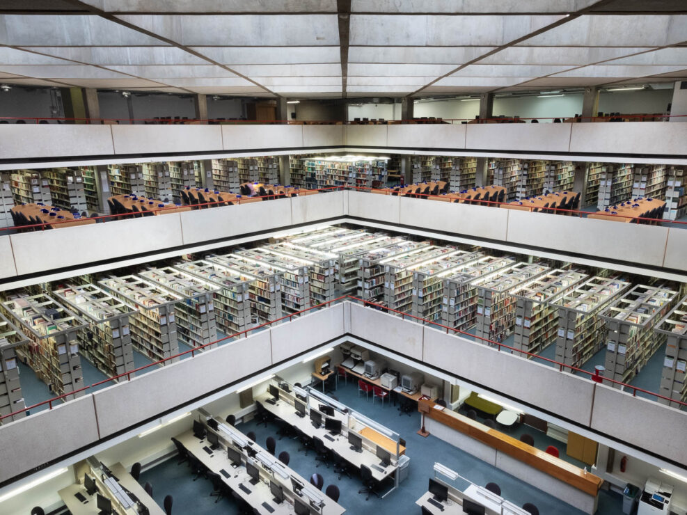 Wide-angle interior shot of a brutalist library. It shows a large atrium with waffle ceiling, three levels of bookshelves stepping down to a floor filled with long tables with computer monitors.