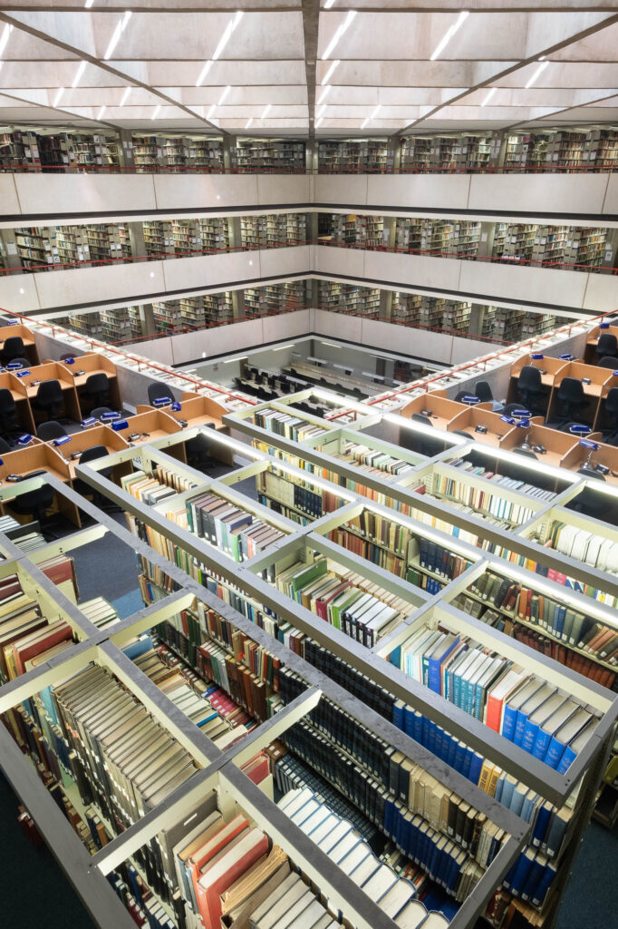 Wide-angle interior shot of a brutalist library. It shows a large atrium with waffle ceiling, three levels of bookshelves stepping down. The forefront is filled with bookshelves viewed from the top.