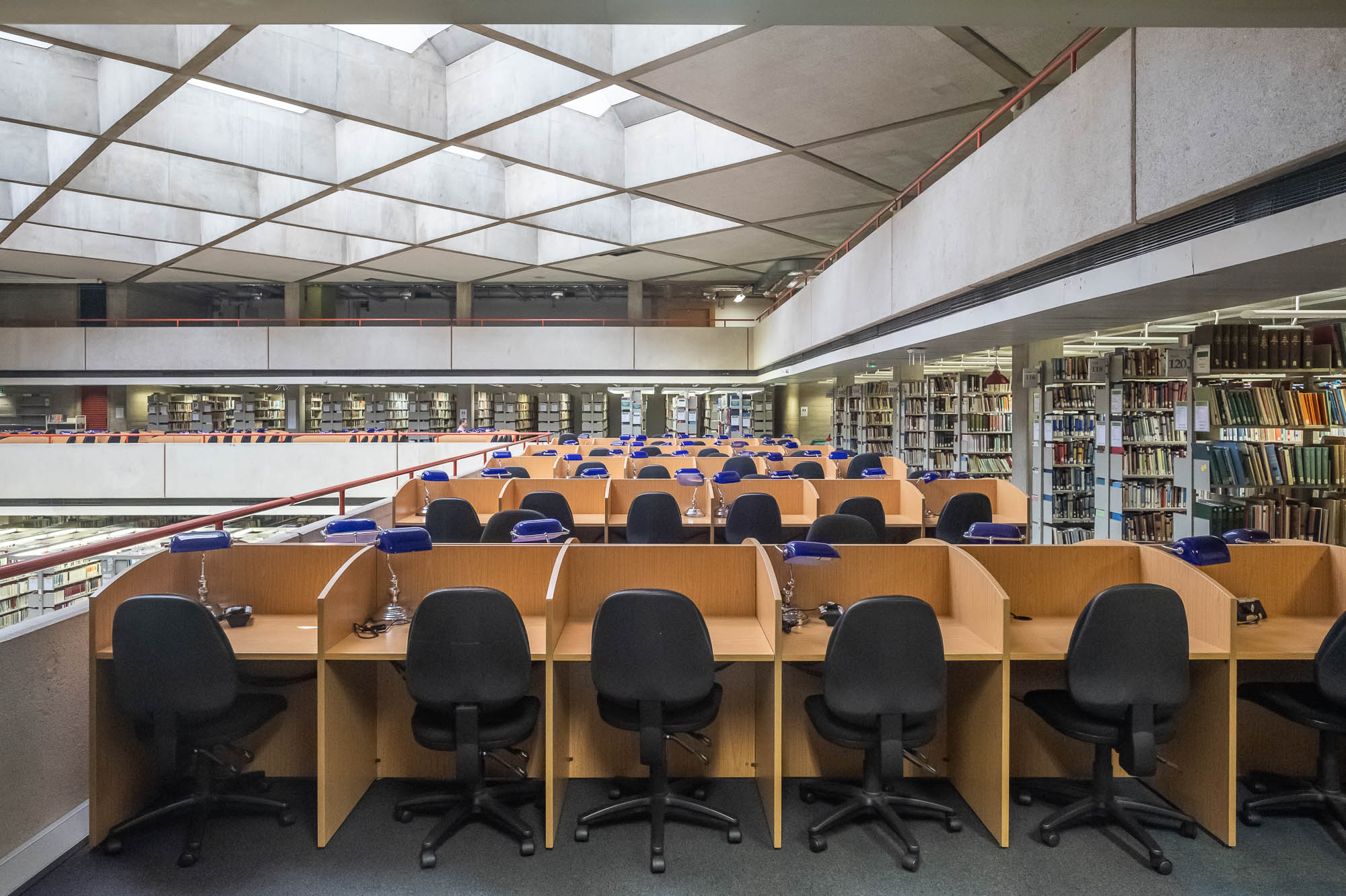 Wide-angle interior shot of a brutalist library. Rows of blond wood cubicles can be seen, each with a modern office chair and library work lamps with blue shades.