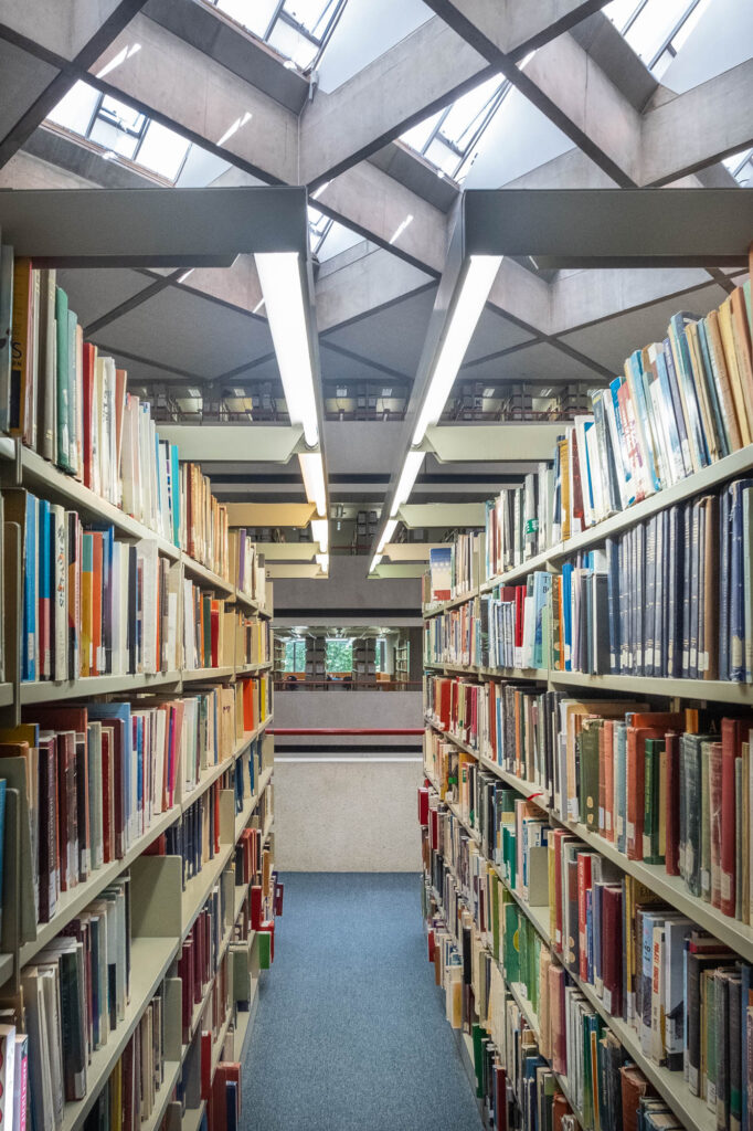 Wide-angle interior shot of a brutalist library. Two rows of bookshelves on either side, the ceiling can be seen between them.