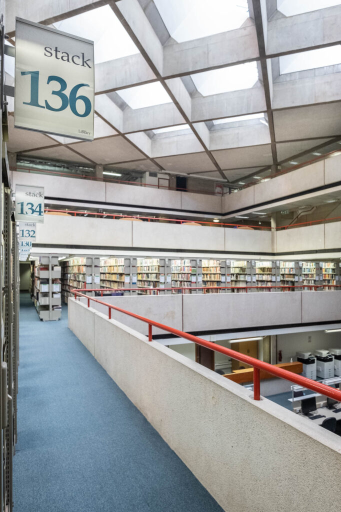 Wide-angle interior shot of a brutalist library. The view is to a level opening to an atrium. To the side are bookshelves with signs reading Stack 136, 134, 132 and so on.