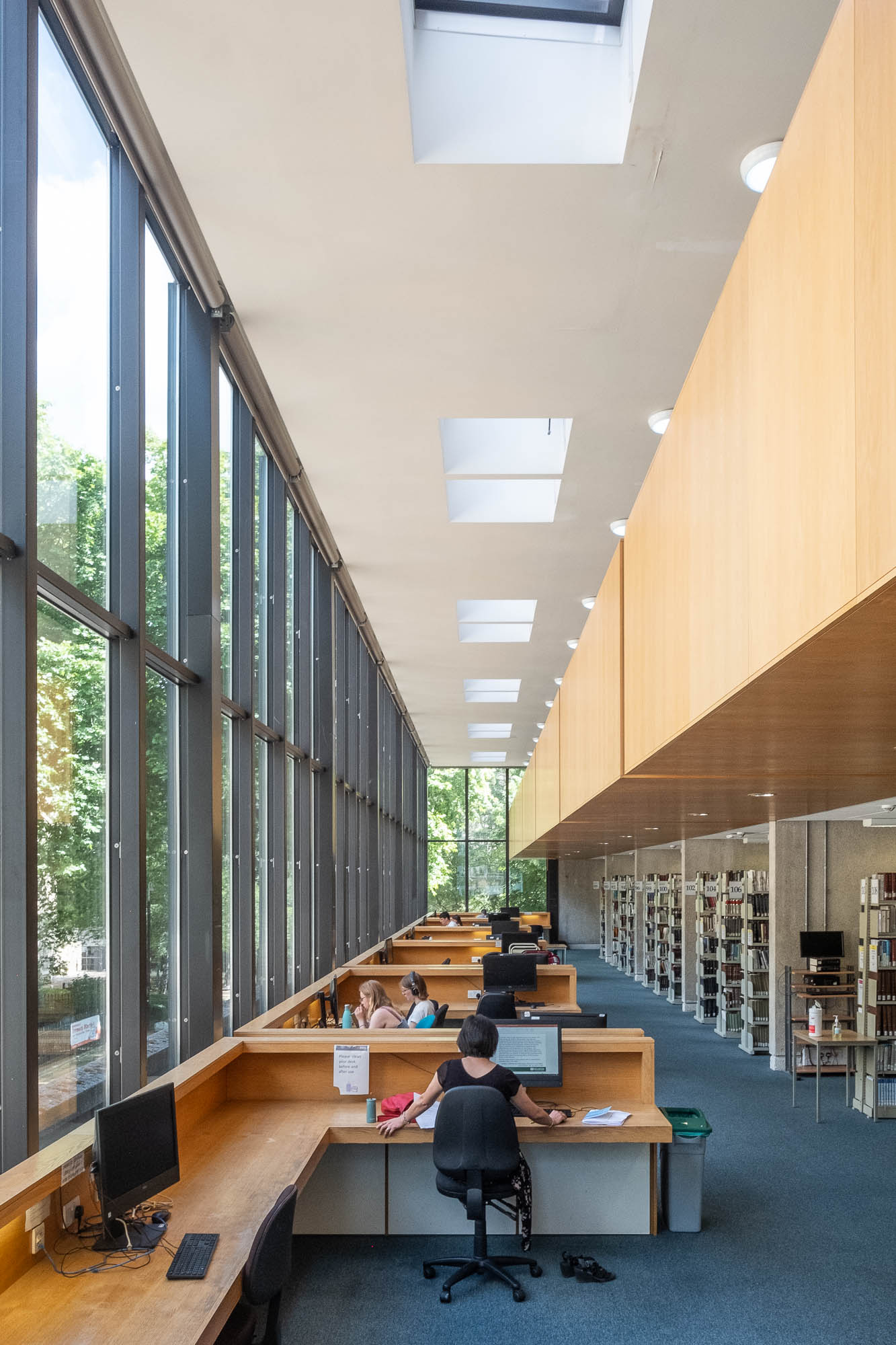 Interior of a modern library reading room with high windows to the left and a mezzanine covered in blond wood to the right. Patrons are seated at large cubicles next to the windows. Bookshelves are seen on the right.