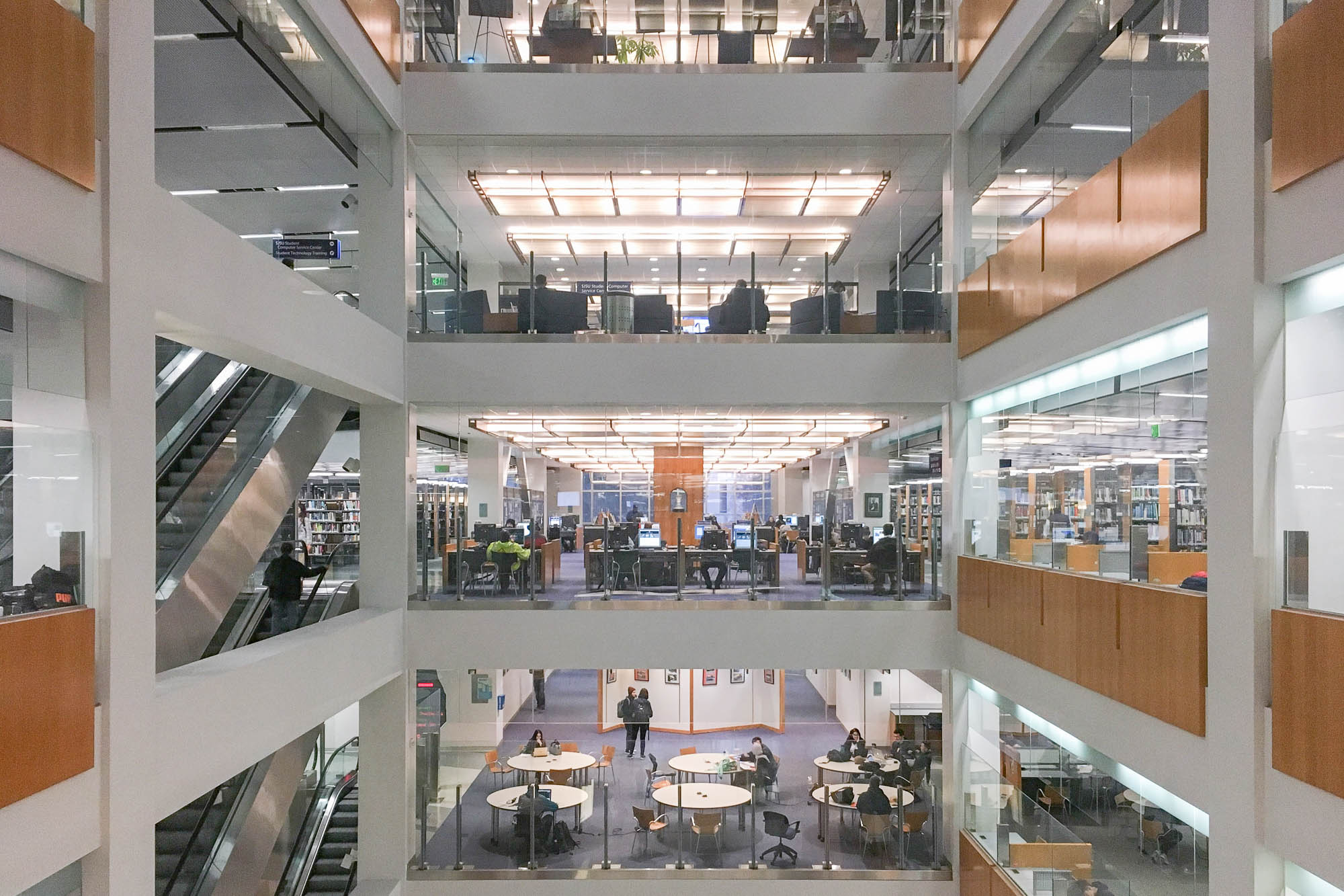 Interior of a postmodern library atrium. We see four levels of the library, with patrons seated at tables, and bookshelves. Escalators are seen to the left.