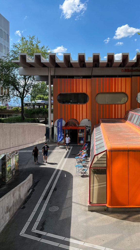 Exterior view of a 1970s pavilion with crenellated roof. The facade is made of vertical stripes of varying shades of orange and pierced with oval windows. In front of the building are bistro tables and chairs, people are walking.