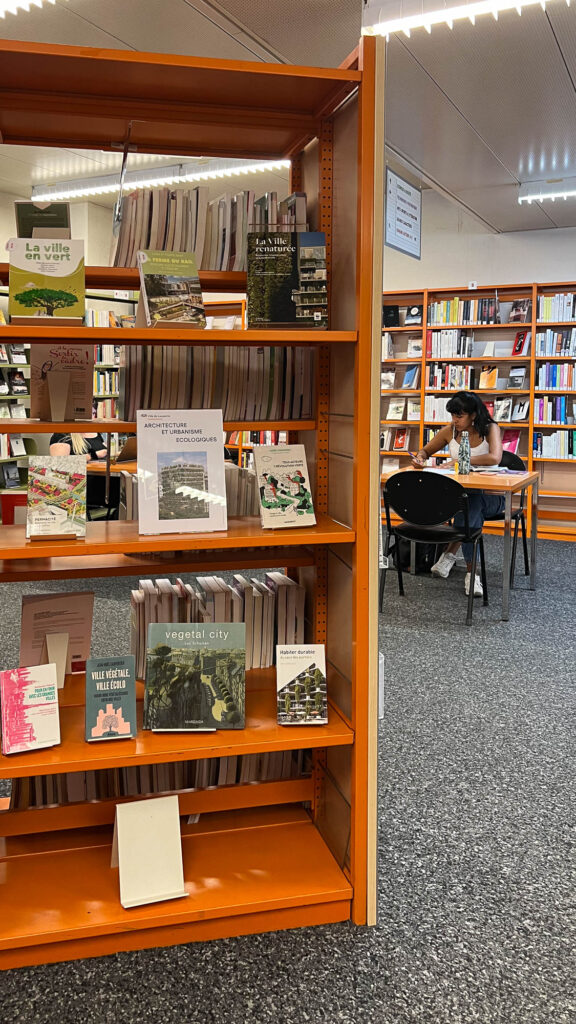 Interior view of a library, with orange metallic shelves. In the back, a person is working on a table.