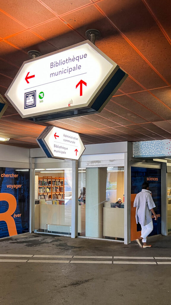 Underground passage with glass frontage, through which a library can be seen. Way-finding signs are suspended from the ceiling, they are an elongated hexagonal shape and show the way to "Bibliothèque municipale" to the left, a train stop towards the back and "Administration communale" towards the front. A person is seen walking.
