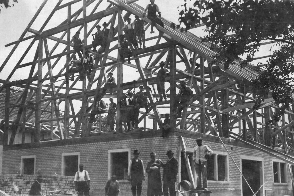 Historical black and white image of a barn being built. It shows a timber structure set on a brick base. Workers are posing on the roof and scaffoldings surrounding the building.
