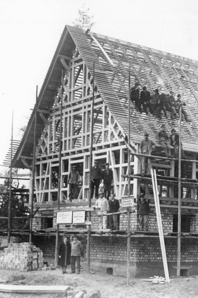 Historical black and white image of a barn being built. It shows a timber structure set on a brick base. Workers are posing on the roof and scaffoldings surrounding the building.
