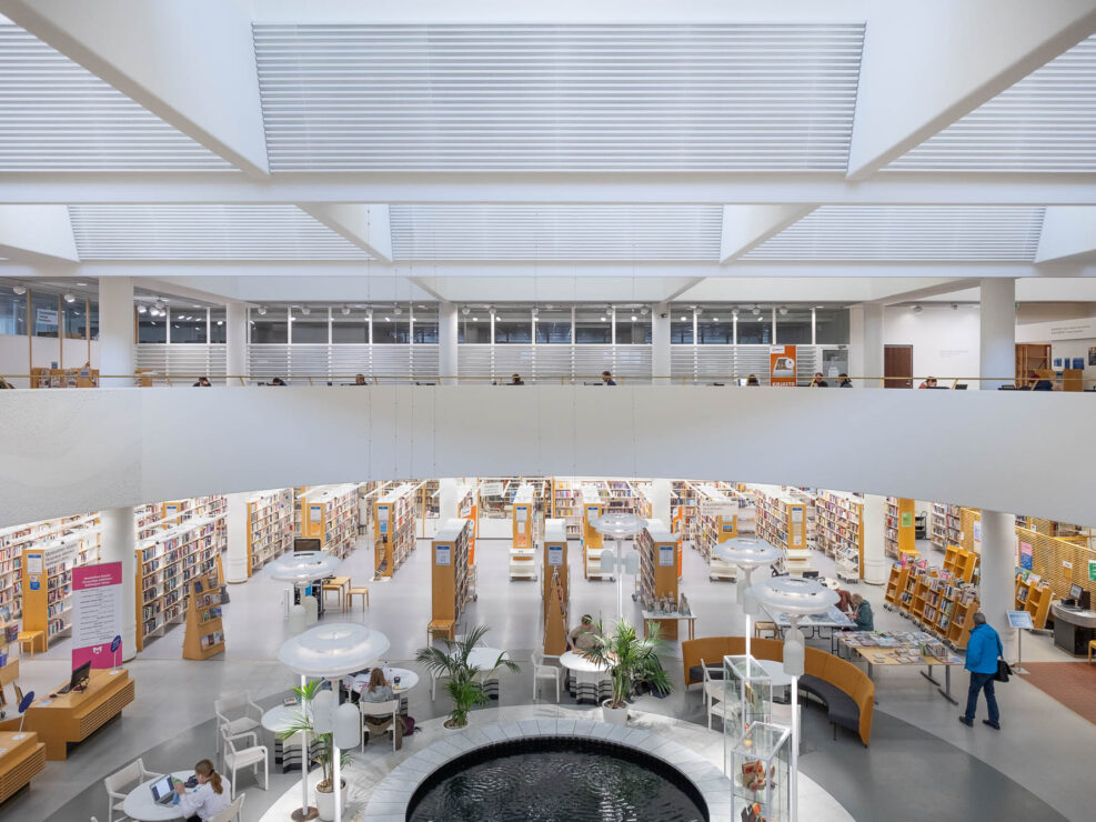 Interior of a modern library, showing a large circular atrium with a fountain in the middle and surrounded by a mezzanine. The photo is taken from the mezzanine. On the bottom floor are rows of bookshelves in blond wood. Round tables, chairs and houseplants are arranged around the fountain.