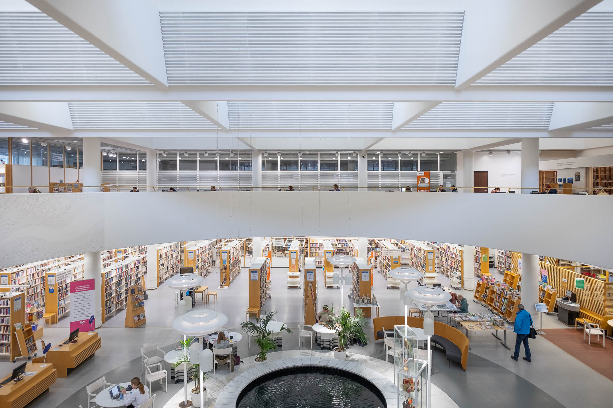 Interior of a modern library, showing a large circular atrium with a fountain in the middle and surrounded by a mezzanine. The photo is taken from the mezzanine. On the bottom floor are rows of bookshelves in blond wood. Round tables, chairs and houseplants are arranged around the fountain.