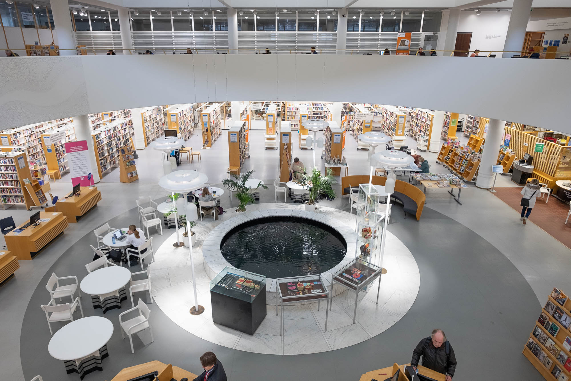 Interior of a modern library, showing a large circular atrium with a fountain in the middle and surrounded by a mezzanine. The photo is taken from the mezzanine. On the bottom floor are rows of bookshelves in blond wood. Round tables, chairs and houseplants are arranged around the fountain.
