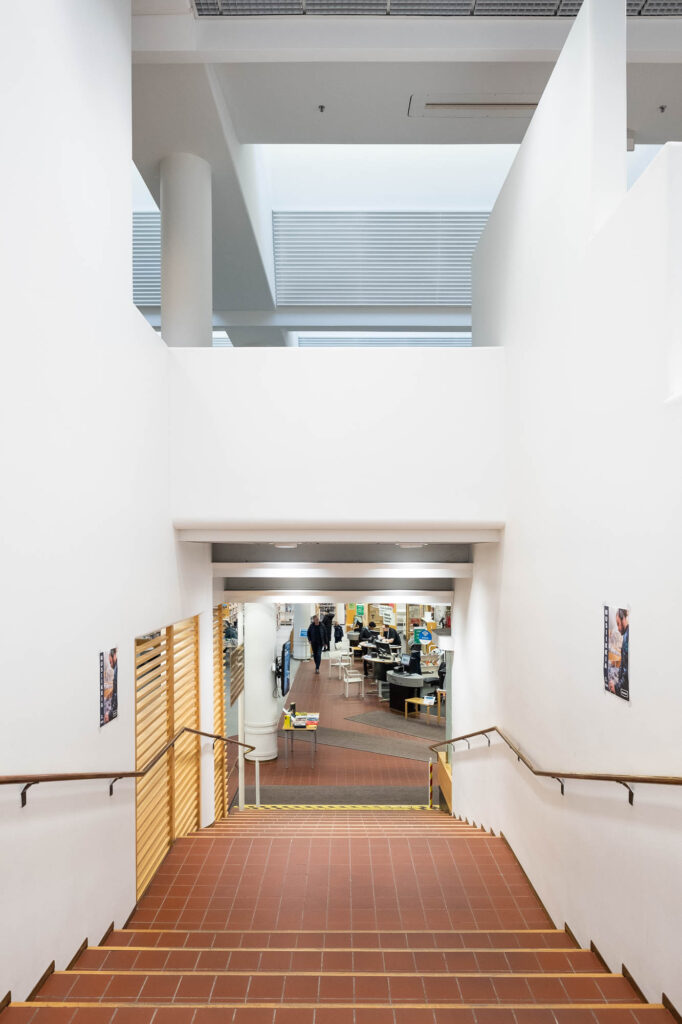 Interior of a modern library, looking down a flight of stairs of red tile with wooden edges. The library interior with tables and patrons can be seen at the bottom.