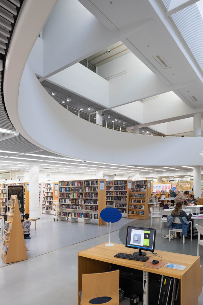 Interior of a modern library, showing the side of a large circular atrium. A workstation is in the foreground and the background shows bookshelves and patrons sitting at round tables.