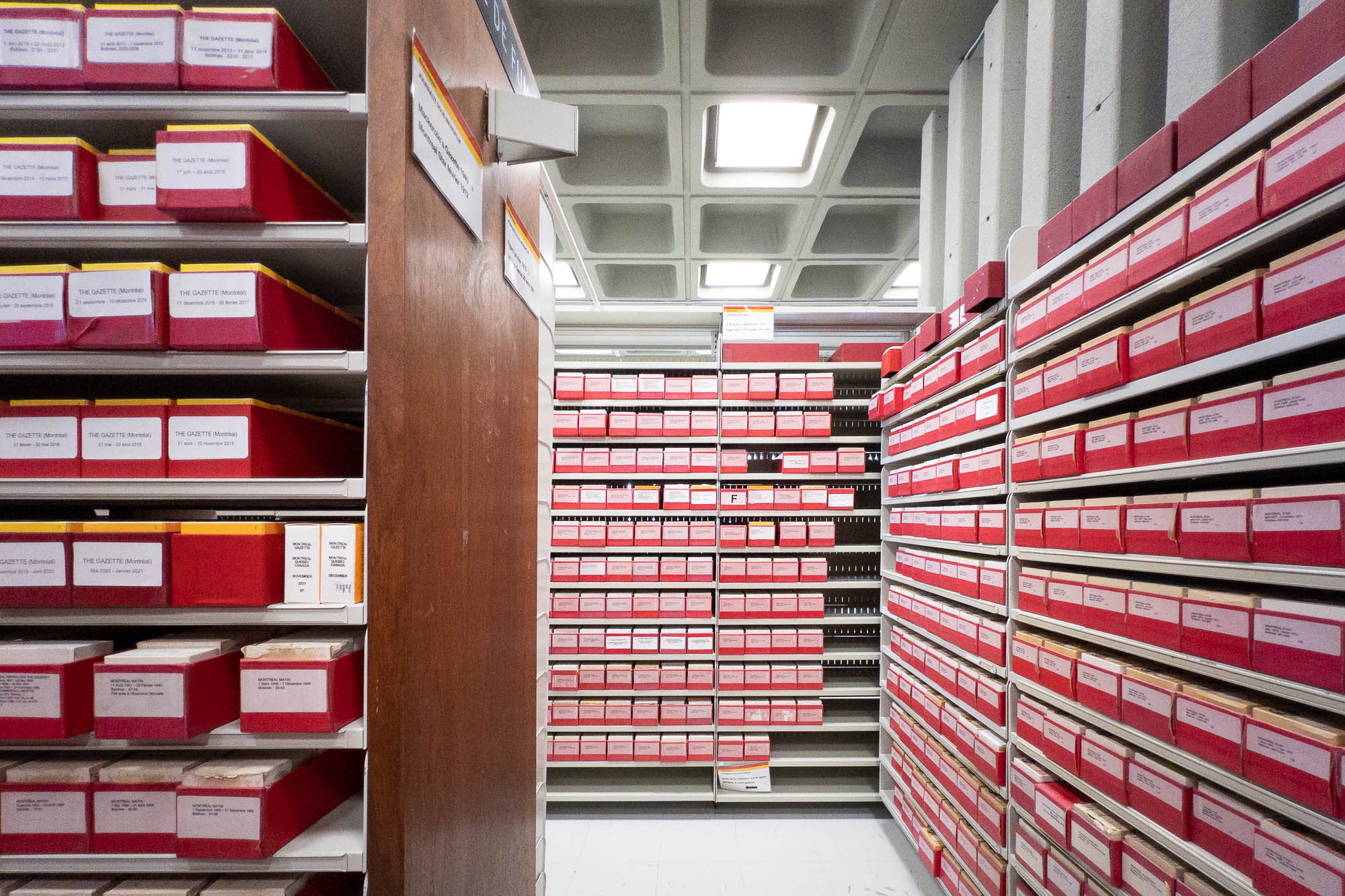 Interior of a brutalist library with waffle ceiling. Walls are occupied by shelves of bright red plastic boxes of microfilms.