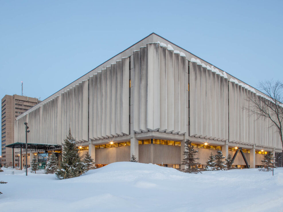 Exterior of a modern library at nightfall. It is a rectangular building with a facade of thin concrete fins. The building is lit with a warm yellow light. There is snow on the ground.