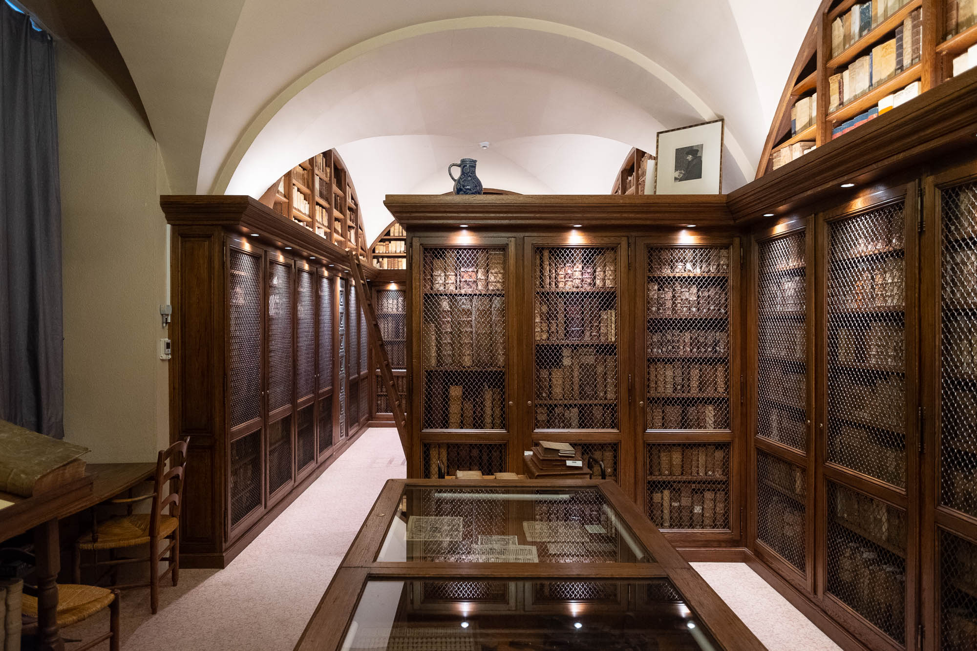 Interior of a rare book library with dark wood shelves filled with precious volumes behind protective netting. The shelves are rounded at the top to fit with the vaulted space. Glass-topped display cases are seen in the foreground.