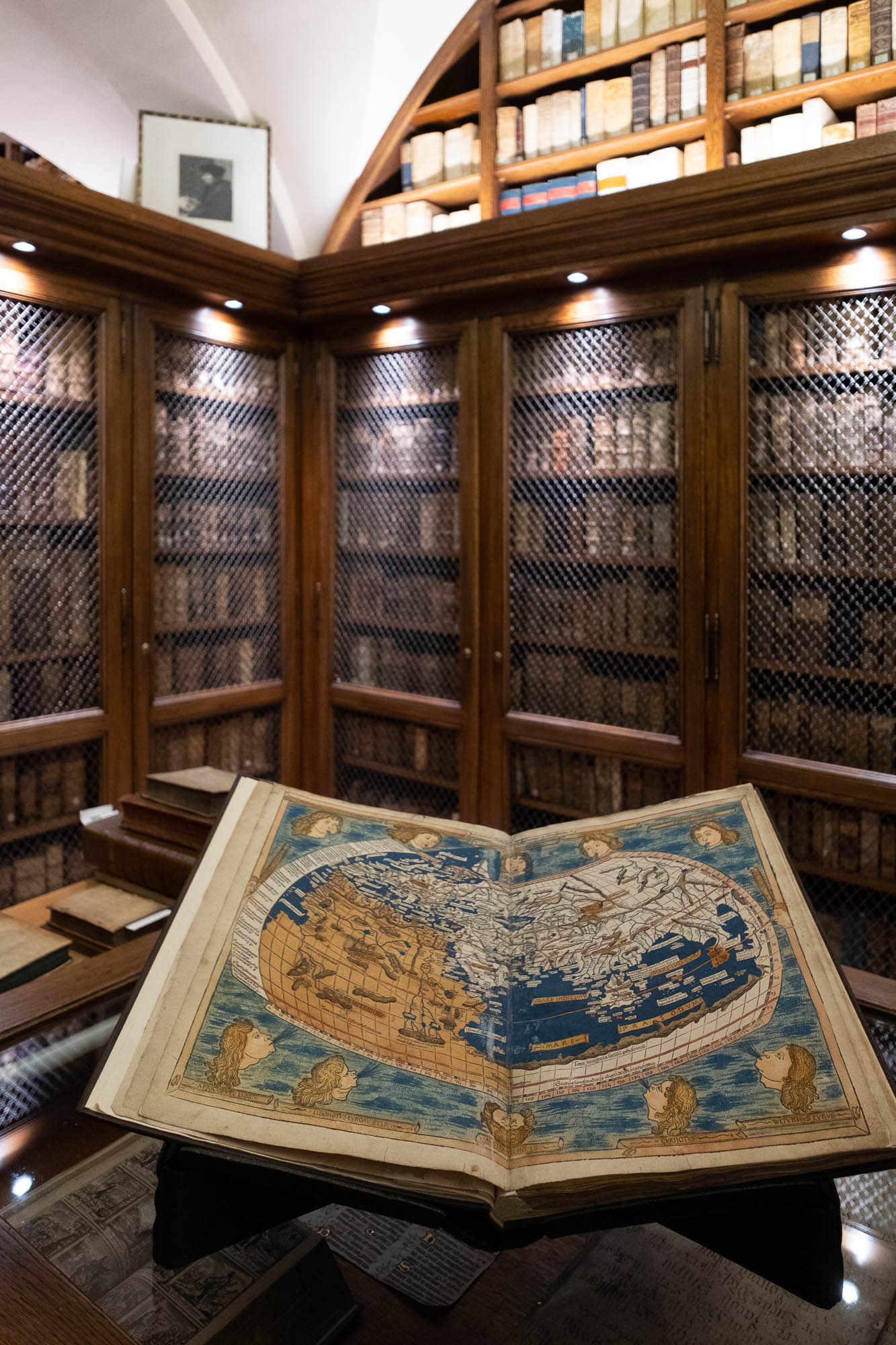 Interior of a rare book library with dark wood shelves filled with precious volumes behind protective netting. An ancient book is seen in the foreground, open to a page representing a map of Europe, Asia and Africa.
