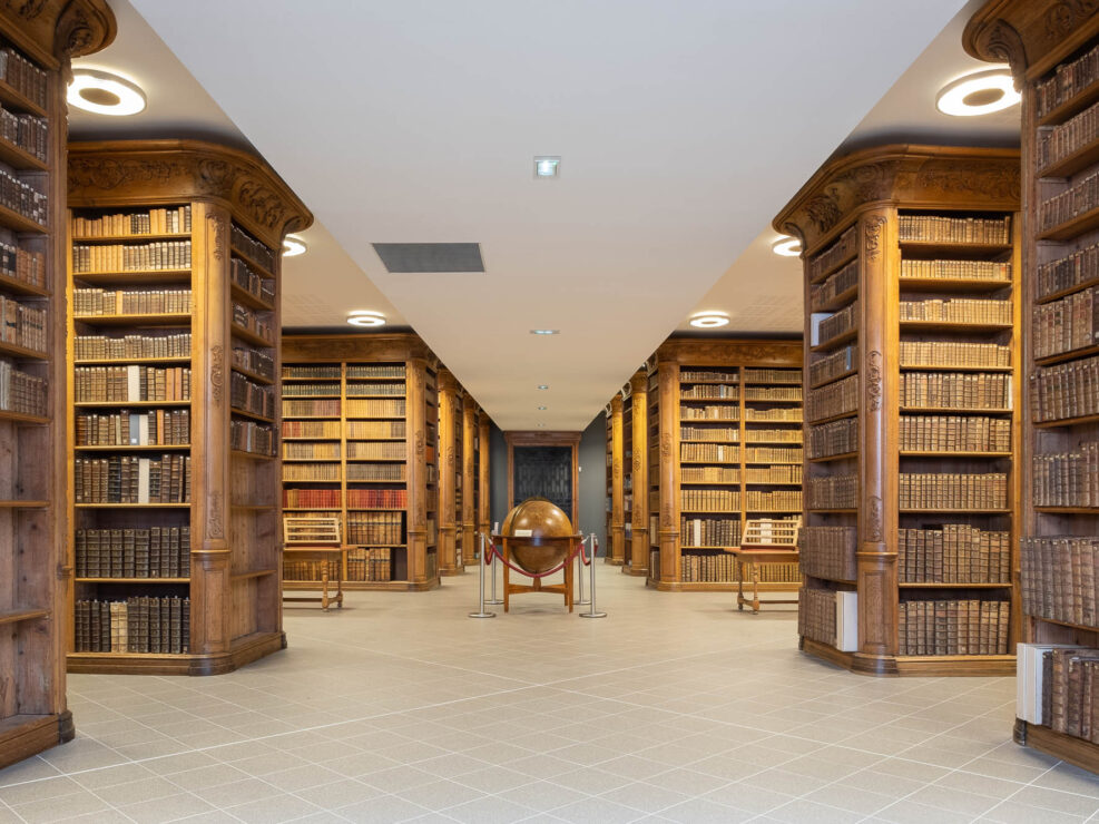Interior view of a patrimonial library with rows of oak bookshelves filled with ancient books on either side and an antique globe in the middle.
