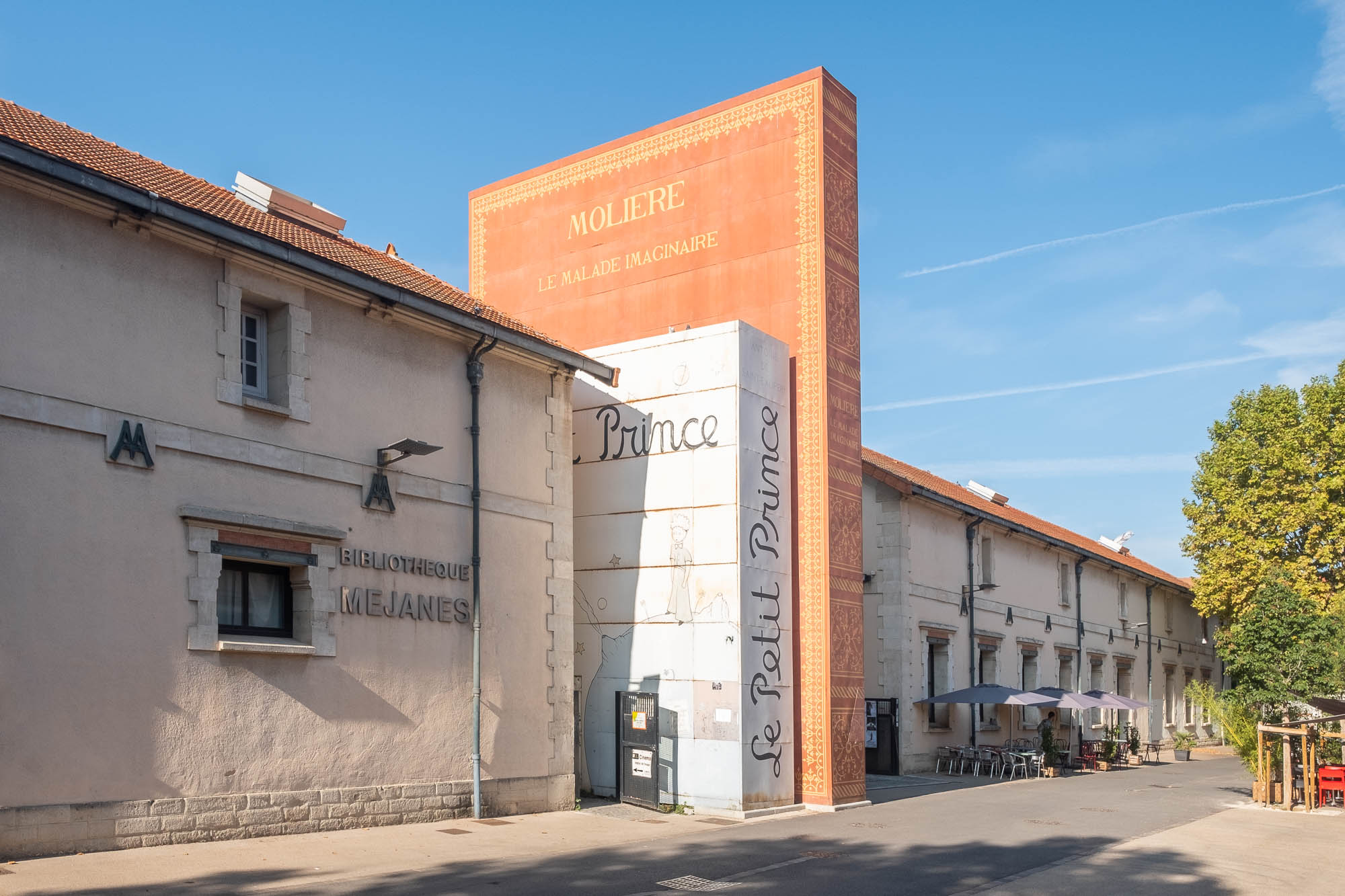 Exterior of a library entrance made with three giant books standing against each other. The titles of the books in sight are "Le Petit Prince" and "Molière: Le malade imaginaire". The words "bibliothèque Méjanes" are printed on the wall in the forefront. At the back, we see a cafe patio with chairs, tables and umbrellas, and large mature trees.