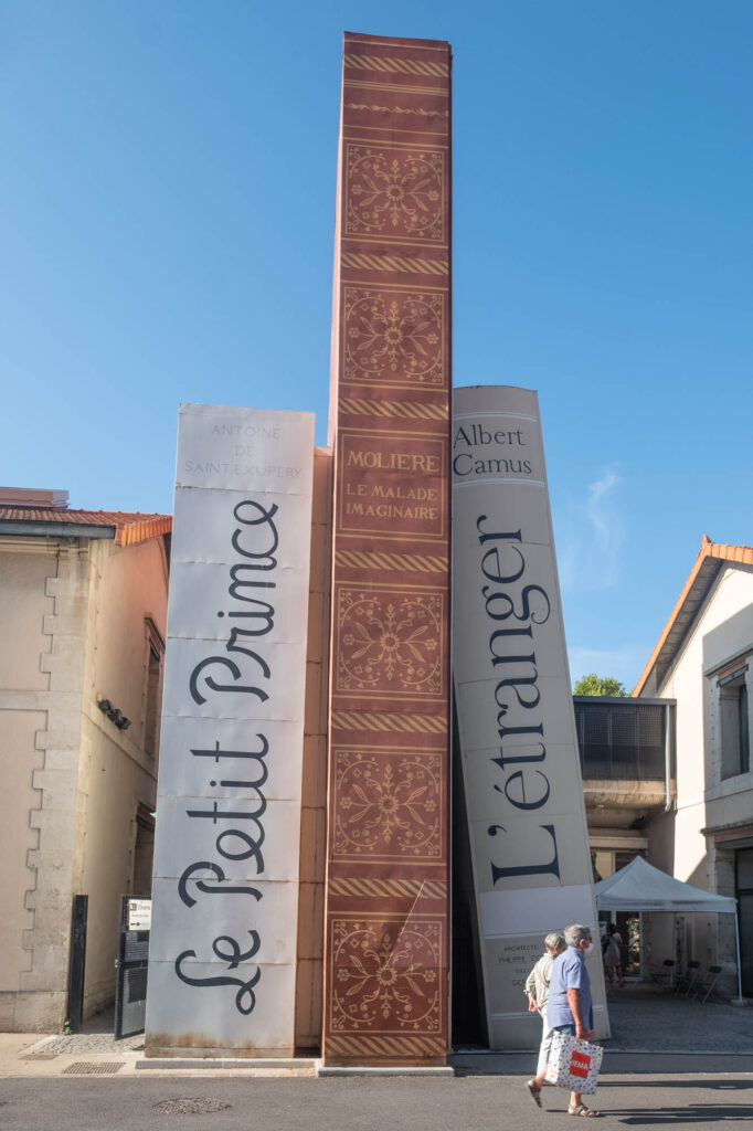 Exterior of a library entrance made with three giant books standing against each other. The titles of the books are "Le Petit Prince", "Molière: Le malade imaginaire" and "Albert Camus: L'étranger".