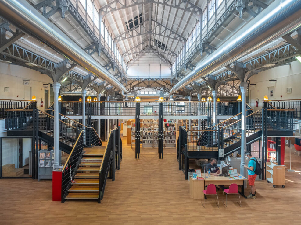 Interior of an industrial space converted into a library with a multistory roof lit by clerestory windows supported on iron beams and poles. A mezzanine crosses the space with stairs on either side. We see bookshelves at the back and a service counter with two people interacting at the front.