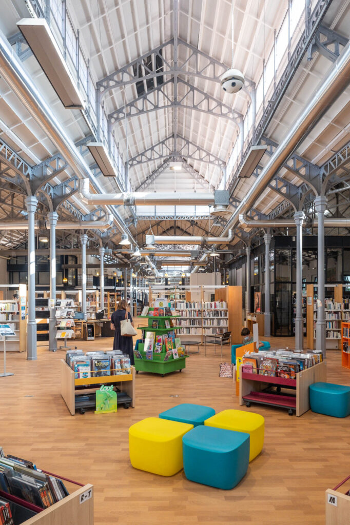 Interior of an industrial space converted into a library with a multistory roof lit by clerestory windows supported on iron beams and poles. Colourful modern library furniture fills the space and patrons are reading.