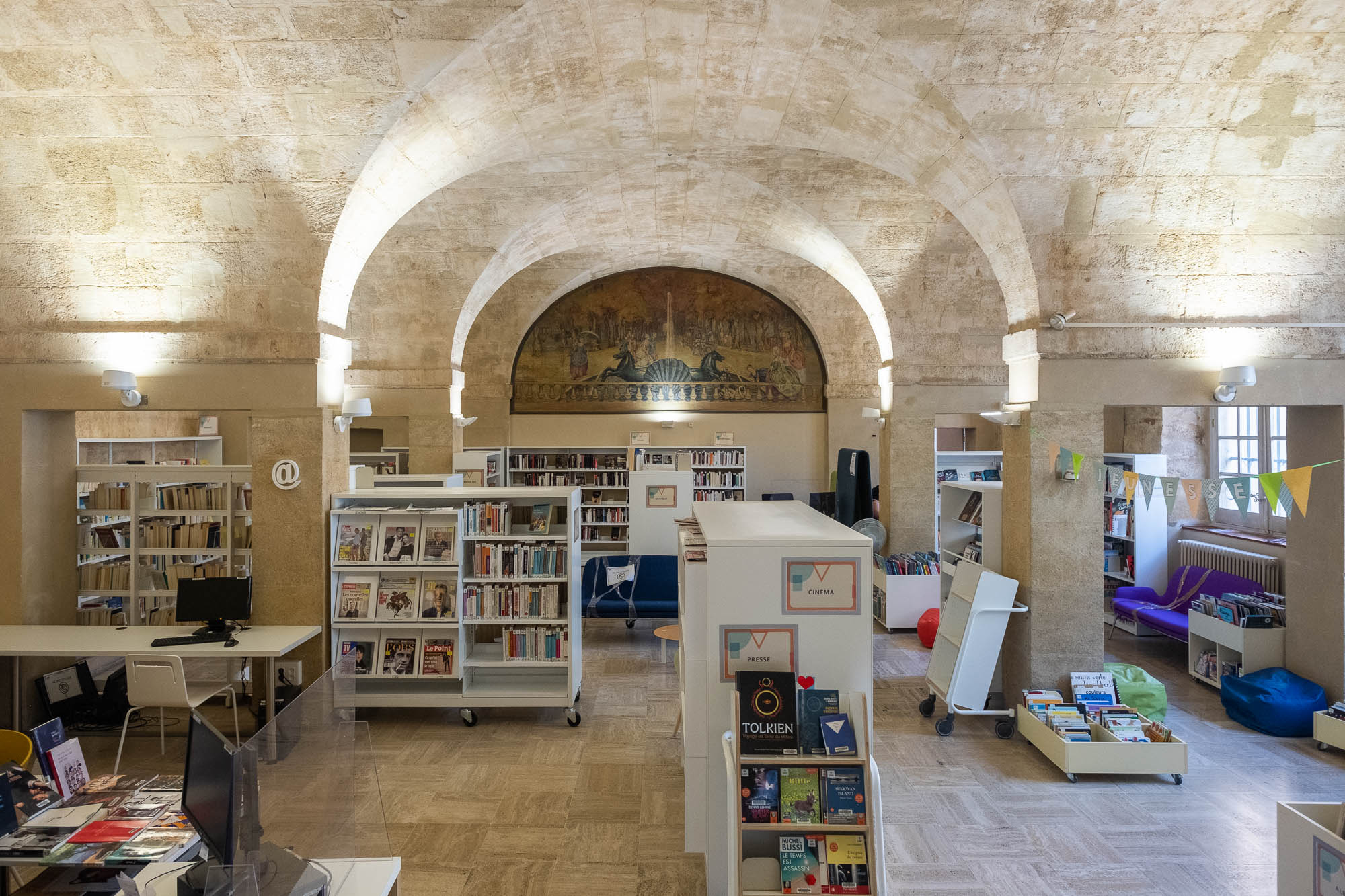 Interior of a library in a stone vaulted space. A fresco showing fountain with two horses and an avenue lined with trees is on the back wall.