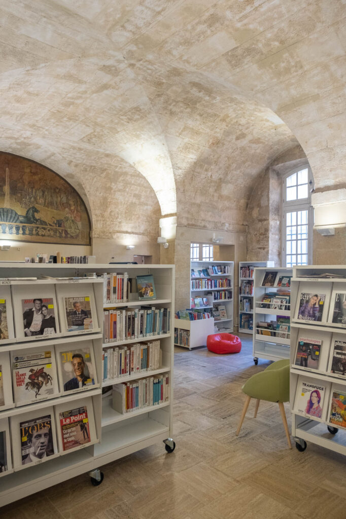 Interior of a library in a stone vaulted space. A fresco showing fountain with two horses and an avenue lined with trees is on the back wall. We see white bookshelves on wheels in the front.