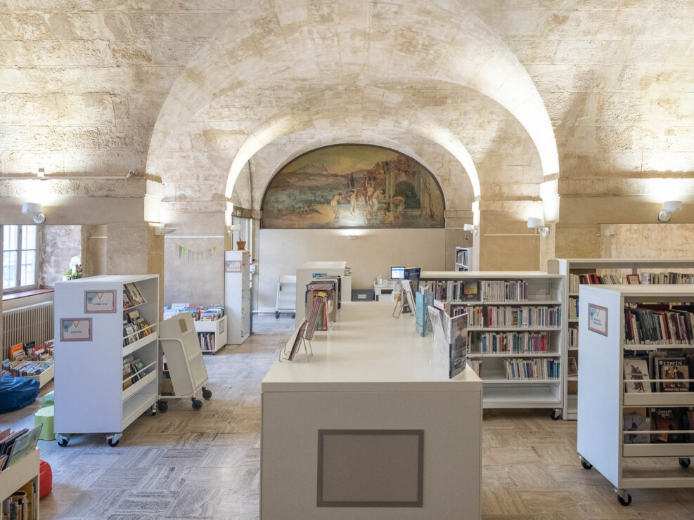 Interior of a library in a stone vaulted space with white modern bookshelves on wheels. A fresco showing a bathing scene is on the back wall.