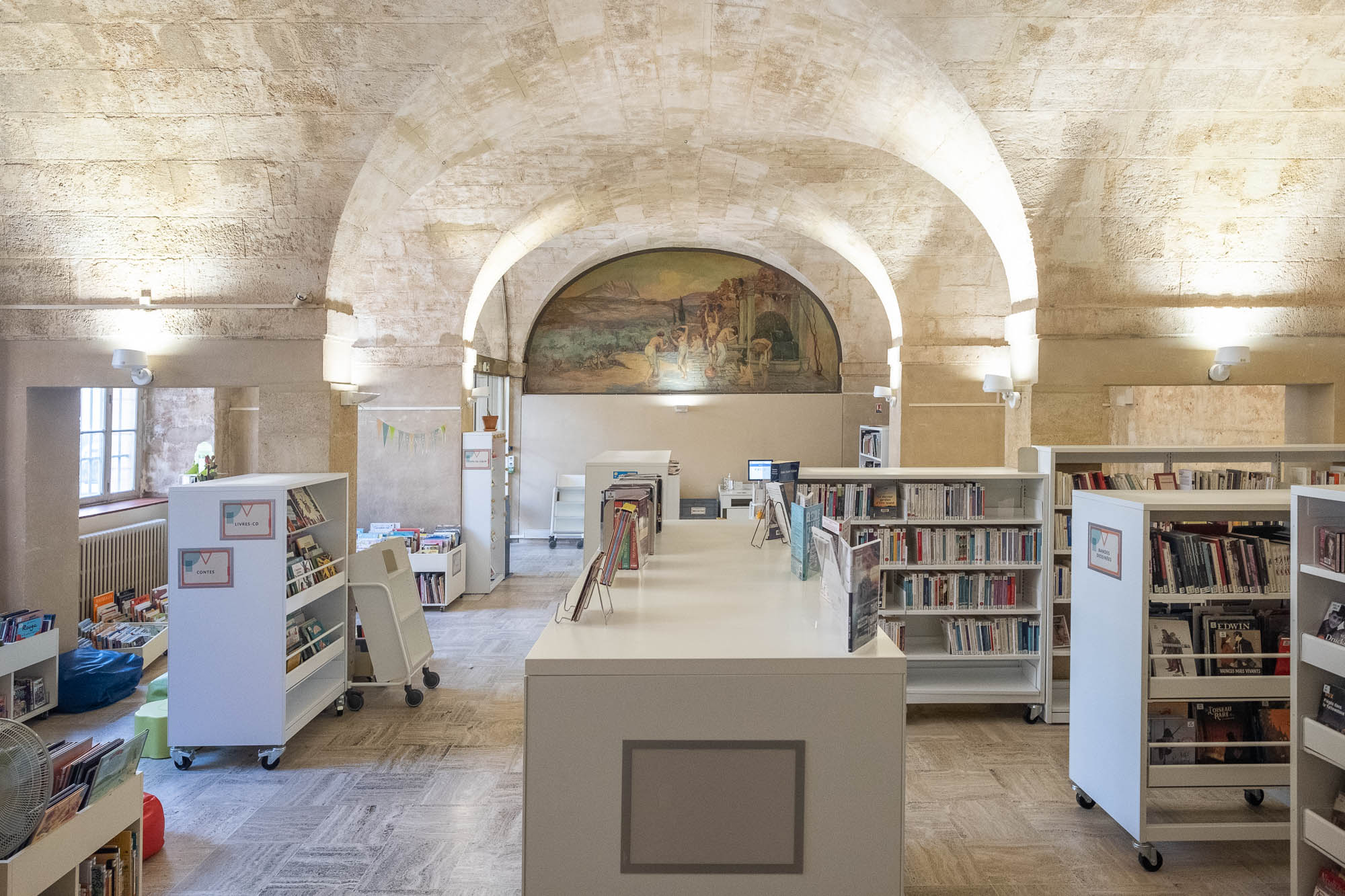 Interior of a library in a stone vaulted space with white modern bookshelves on wheels. A fresco showing a bathing scene is on the back wall.