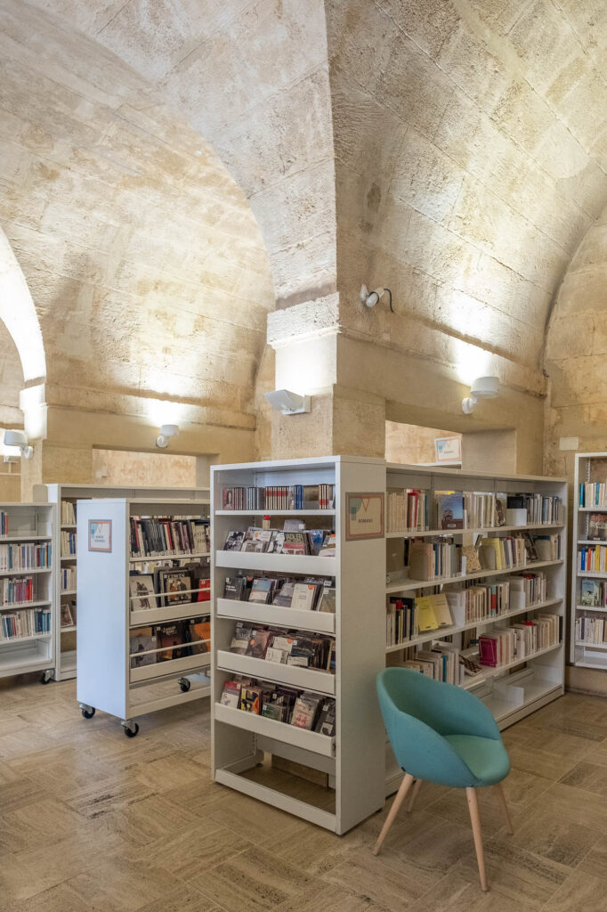 Interior of a library in a stone vaulted space lit from below with indirect light. There are modern white bookshelves and an upholstered chair.