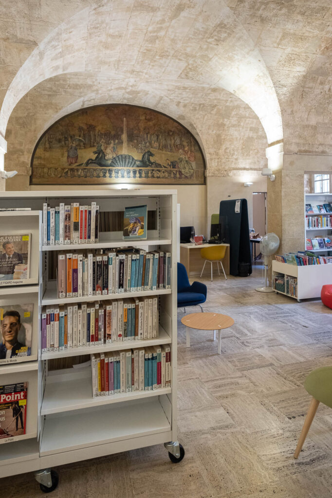 Interior of a library in a stone vaulted space. A fresco showing fountain with two horses and an avenue lined with trees is on the back wall. We see white bookshelves on wheels in the front.