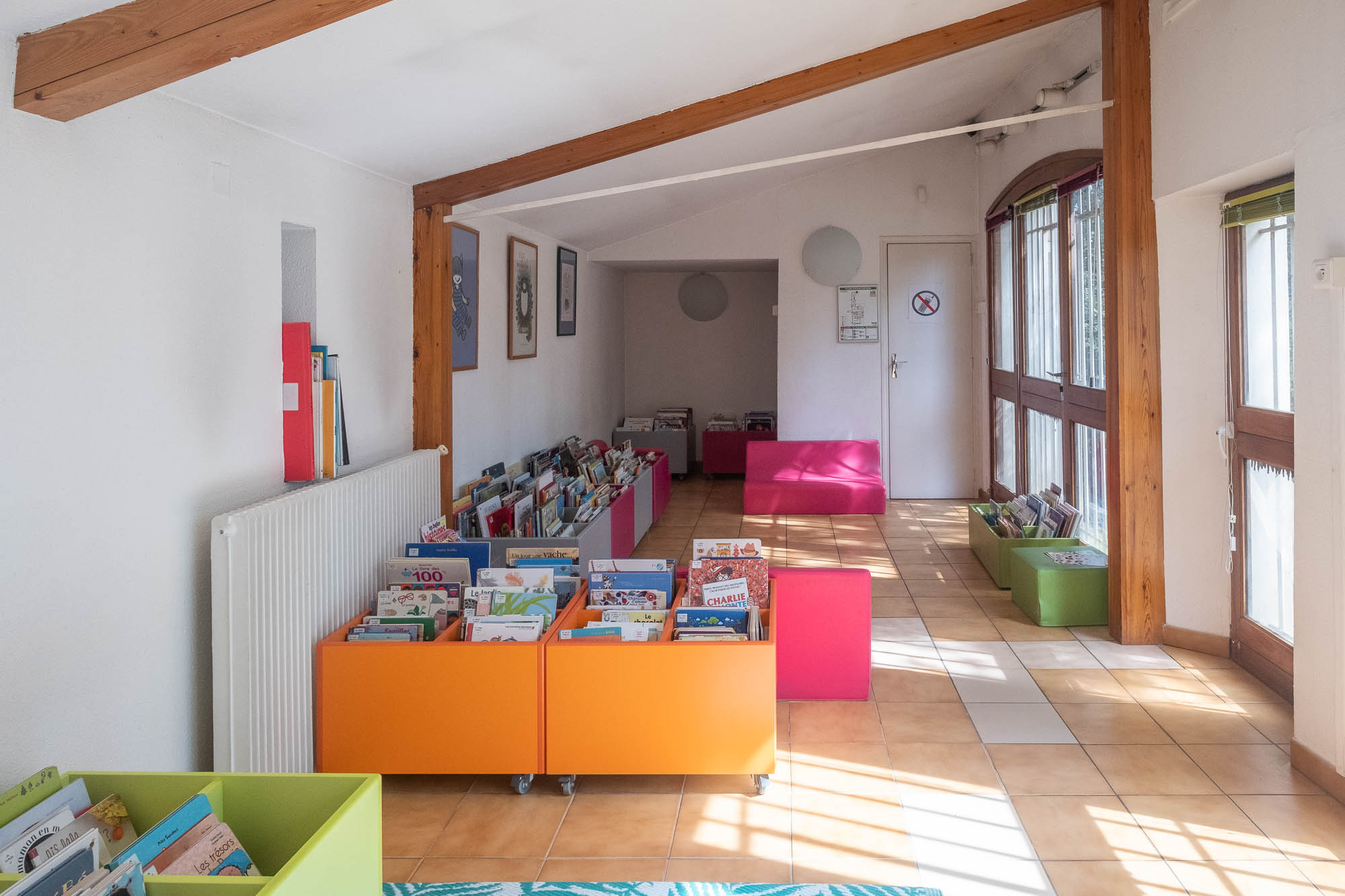 Interior of an old bastide house transformed into a library, showing a room with a slanted roof with exposed wood beam and rolling boxes full of children's books on the tiled floor.
