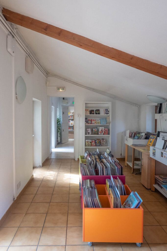 Interior of an old bastide house transformed into a library, showing a room with a slanted roof with exposed wood beam and rolling boxes full of children's books on the tiled floor.