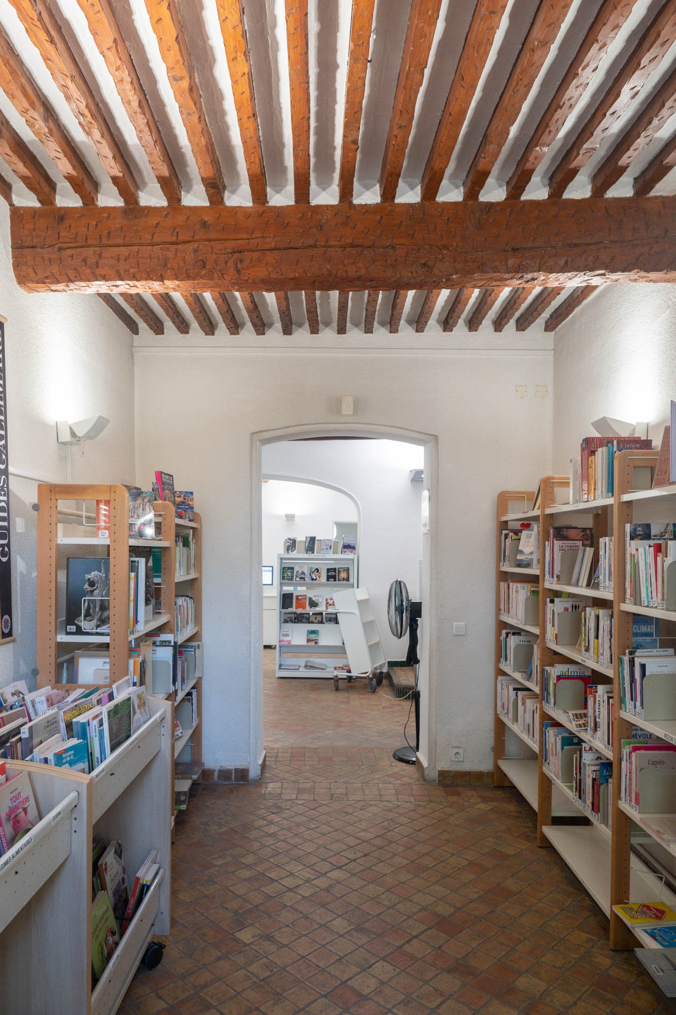 Interior of an old bastide house transformed into a library with wooden beams showing on the ceiling, arched doorways and modern wooden shelves along the walls.