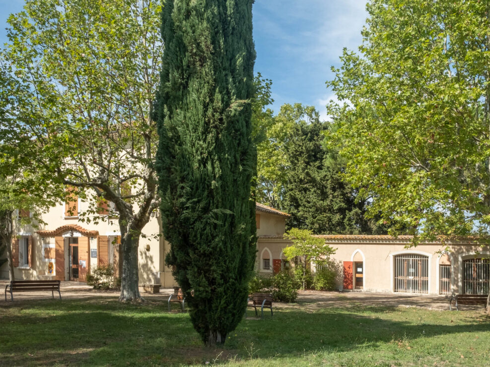 Exterior of a traditional French bastide house in a lush green park, with trees in the foreground.