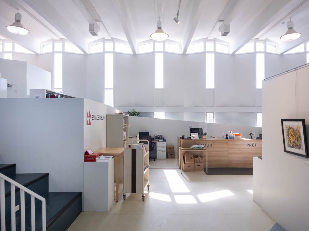 Interior of a modern library, with a service counter at the back with the words "Retour" and "Prêt". To the left is a set of stairs leading to an elevated area with bookshelves. The words "Espace biblio" are written on a partition wall. To the right, an art print representing flowers hangs from a wall. The back wall is structured in a zigzag pattern, with T-shaped windows in each fold.
