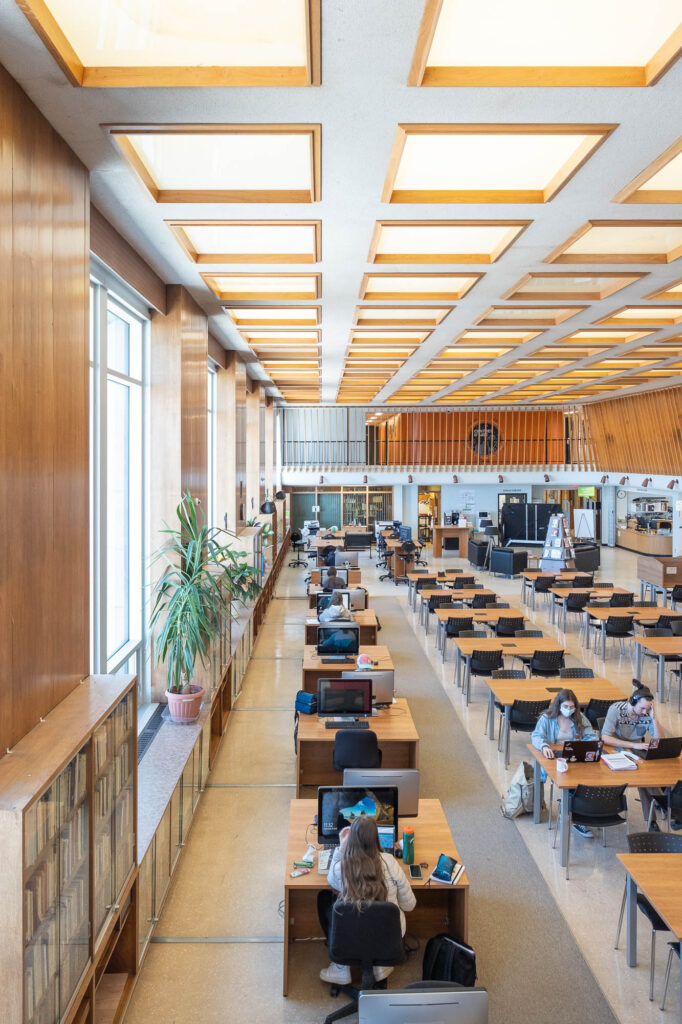 Interior of a modern library with wood-panelled walls. It is a large room with rows of tables and large windows to the left. A large houseplant sits on the window ledge. Students are working at the tables and computer workstations.