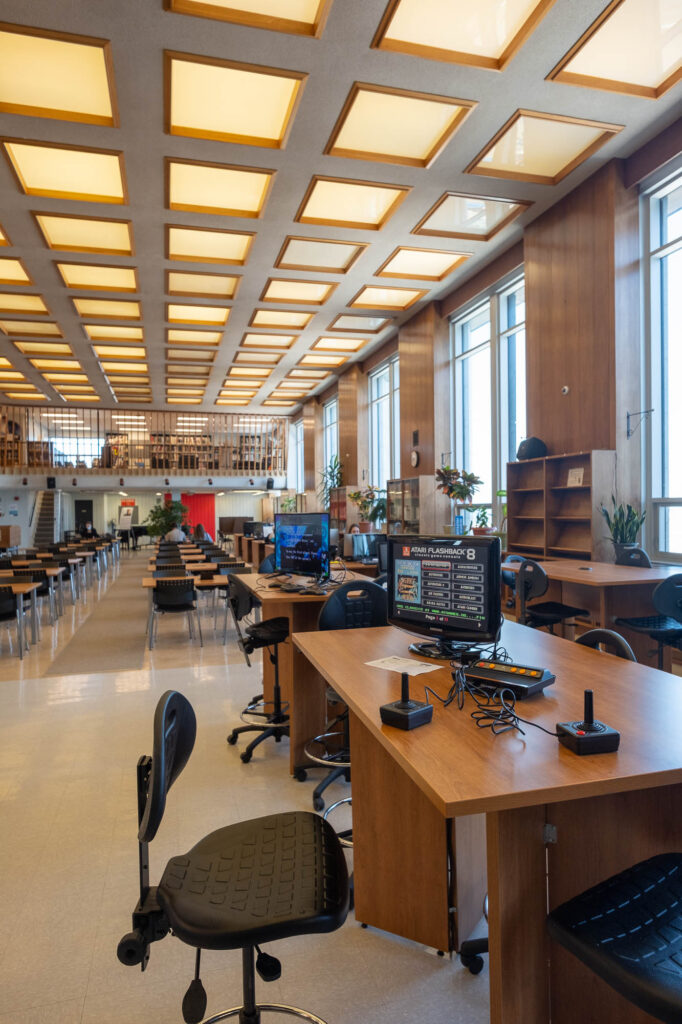 Interior of a modern library with wood-panelled walls. It is a large room with rows of tables and large windows to the right. The ceiling is made out of square light boxes. A computer with a gaming console sits on a table at the front.
