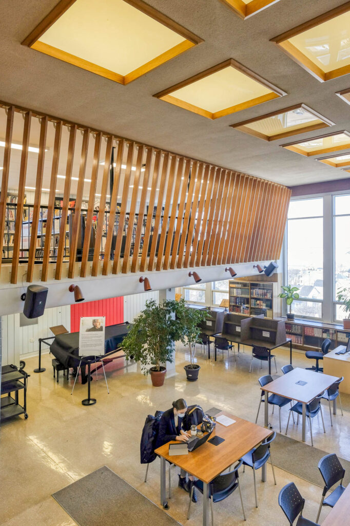 Interior of a modern library. The image is taken from a mezzanine towards a reading room below, where a person seated at a table works on a laptop computer.