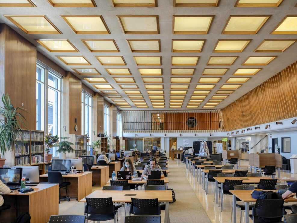 Interior of a modern library with wood-panelled walls. It is a large room with rows of tables and large windows to the left. Students are working at the tables and computer workstations.