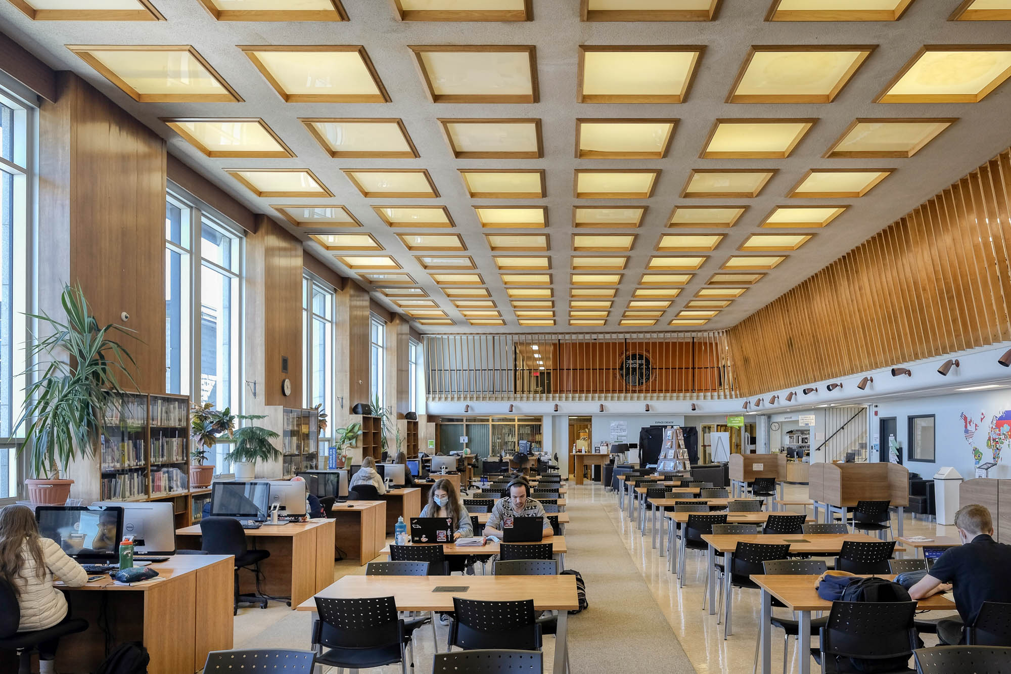 Interior of a modern library with wood-panelled walls. It is a large room with rows of tables and large windows to the left. Students are working at the tables and computer workstations.