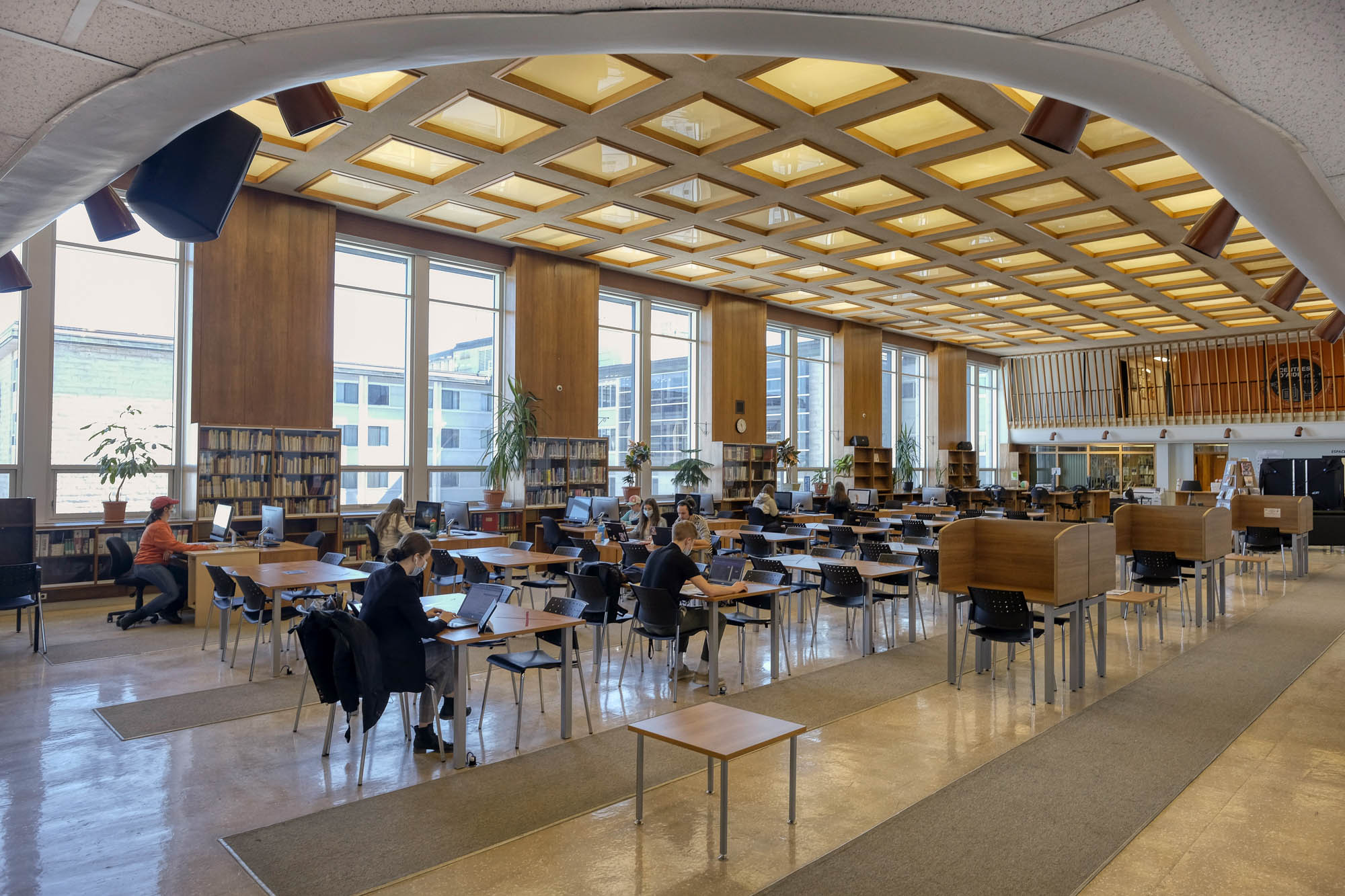 Interior of a modern library with wood-panelled walls. It is a large room with rows of tables and large windows on the back wall. Students are working at the tables.