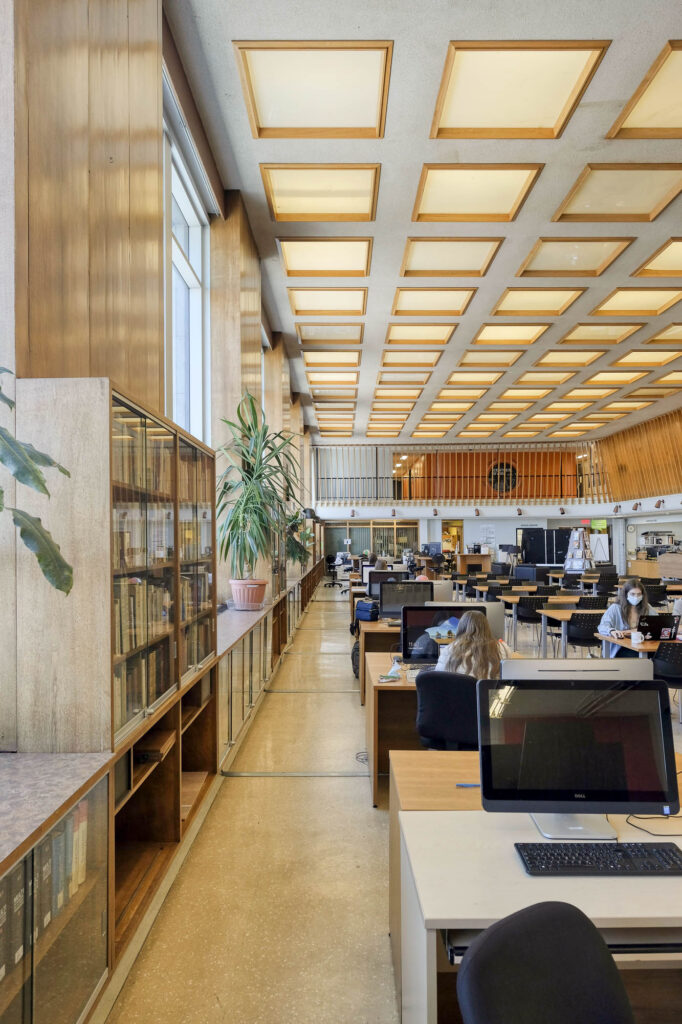 Interior of a modern library with wood-panelled walls. It is a large room with rows of tables and large windows to the left. A large houseplant sits on the window ledge. Students are working at the tables and computer workstations.