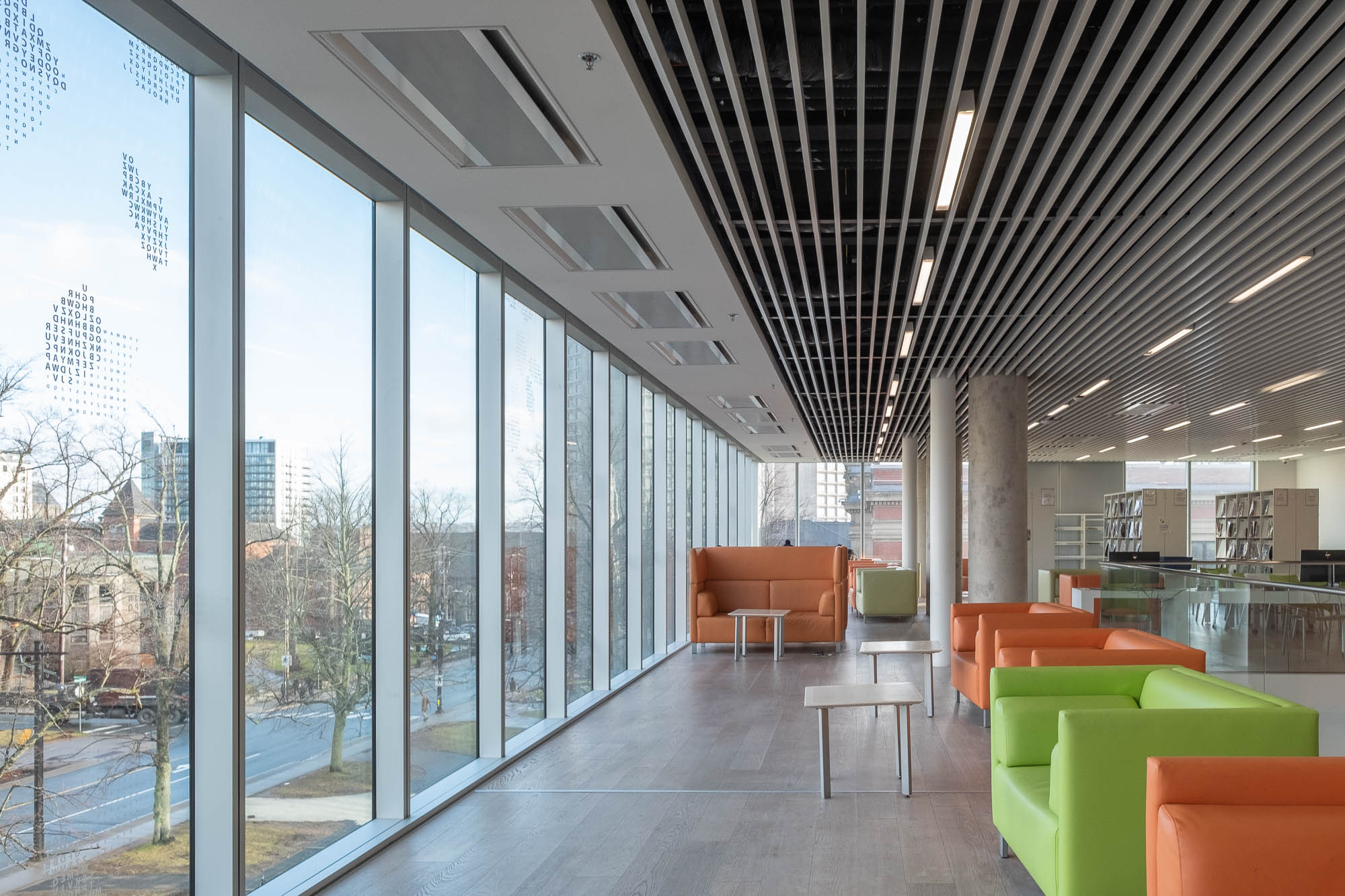Interior of a contemporary library, with a glass facade to the left opening to a quiet street. Colourful couches face the window to the right.