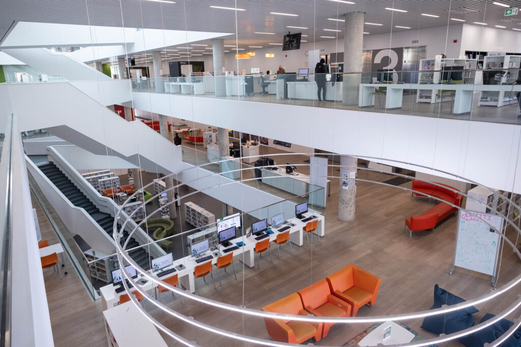 Interior of a contemporary library, with criss-crossing stairs in an atrium. In the foreground are colourful couches, with a large circular light hanging above them.