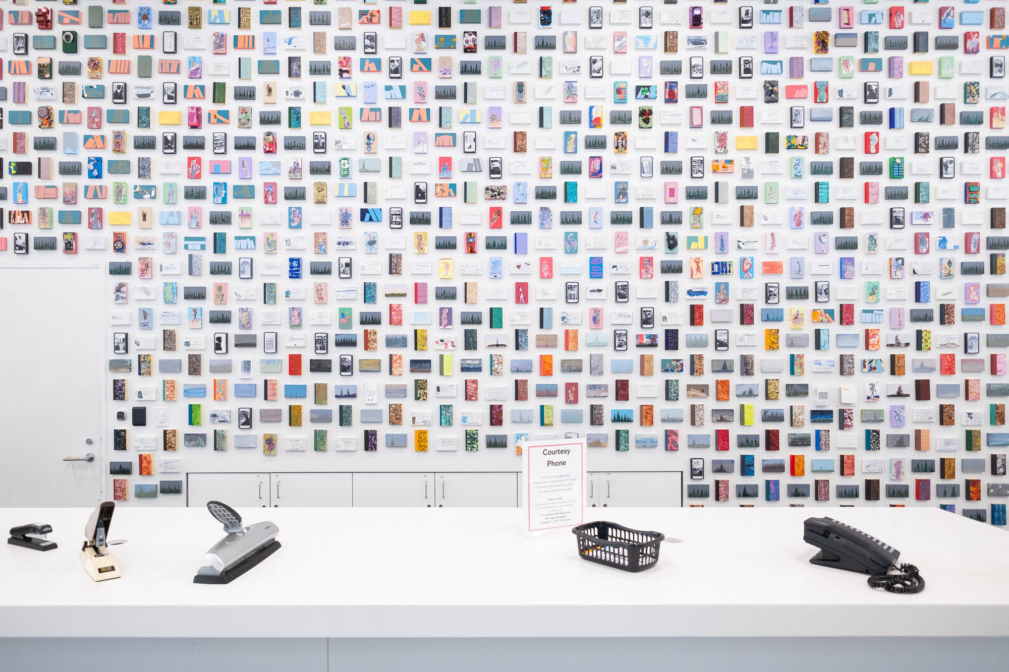 Interior of a contemporary library, showing artwork on a wall, made out of a great number of small colourful rectangles, on which motifs are painted. Some have found objects glued to them. In the foreground is a table with library paraphernalia: staplers, phone, box of pencils.