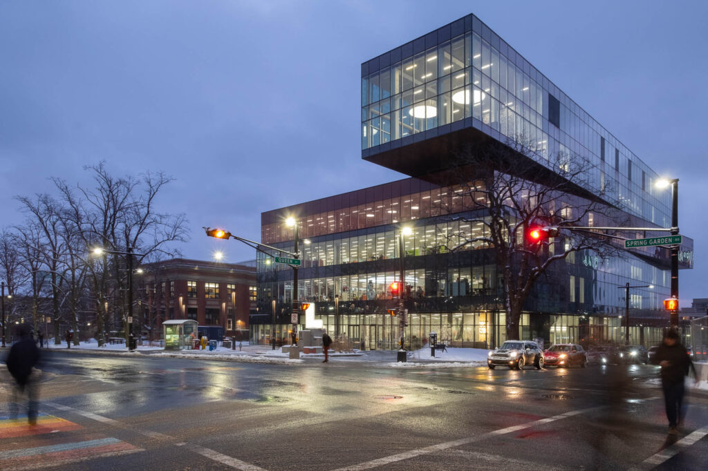 Exterior of a contemporary library at nightfall. Each level is slightly offset from the others, resembling a stack of boxes. The top floor is cantilevered over the plaza below. The library is brightly lit from the inside.