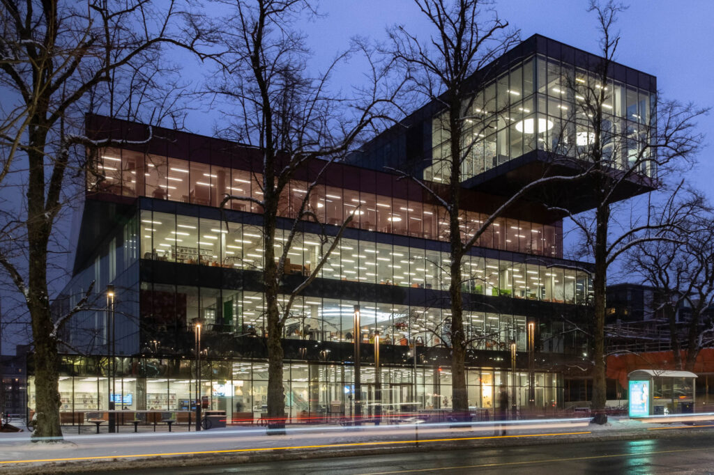Exterior of a contemporary library at nightfall. Each level is slightly offset from the others, resembling a stack of boxes. The library is brightly lit from the inside.